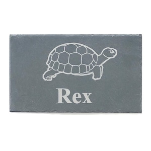 Pode incluir: Uma tábua de queijo retangular de ardósia cinza com um desenho de tartaruga gravado em branco e o nome "Rex" em uma fonte serif. A tartaruga é retratada em perfil, com sua carapaça e pernas detalhadas. A tábua está em um fundo branco.