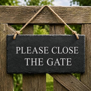A dark grey rectangular slate plaque hangs by a jute rope on a wooden gate, featuring the engraved text PLEASE CLOSE THE GATE.