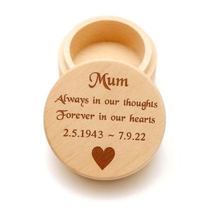 Puede incluir: Una caja redonda de madera clara con tapa. La tapa está grabada con la palabra "Mum", las frases "Always in our thoughts, Forever in our hearts", las fechas 2.5.1943 - 7.9.22 y un símbolo de corazón. La caja está abierta, revelando el interior.