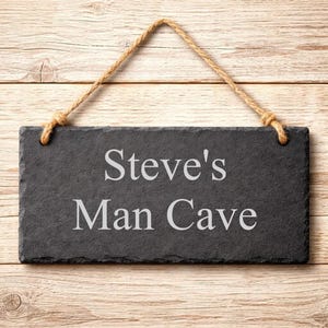 A rectangular dark grey slate plaque hangs by a jute rope on a wood background, engraved with the words Steve's Man Cave.