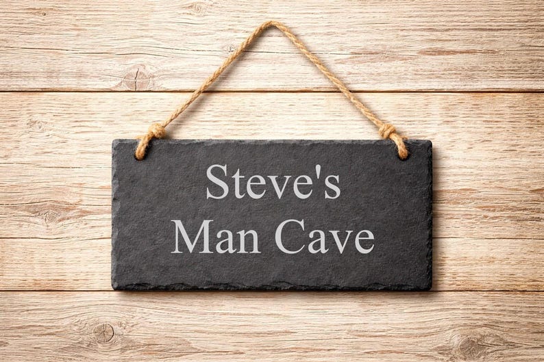 A rectangular dark grey slate plaque hangs by a jute rope on a wood background, engraved with the words Steve's Man Cave.