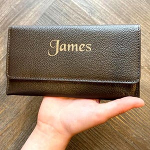 May include: A dark brown leather wallet with the name "James" embossed in gold lettering. The wallet has a textured finish and a flap closure. The wallet is held in a person's hand.
