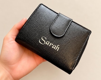 May include: A black bi-fold wallet with a textured, pebbled surface and a snap-button closure flap. The name Sarah is printed in a white, elegant serif font on the front center of the wallet, which features a compact, rectangular shape.