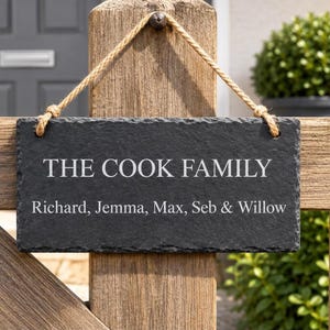 A dark rectangular slate plaque hangs by a twine rope on a rustic wooden gate, engraved with the text THE COOK FAMILY and Richard, Jemma, Max, Seb & Willow.