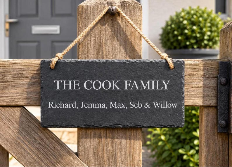 A dark rectangular slate plaque hangs by a twine rope on a rustic wooden gate, engraved with the text THE COOK FAMILY and Richard, Jemma, Max, Seb & Willow.