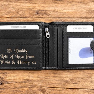 May include: A black bi-fold wallet featuring multiple card slots, a central zippered compartment, and a clear ID window. The interior is engraved with the text: To Daddy Lots of Love from Olivia & Harry xx. The wallet includes a snap button closure.