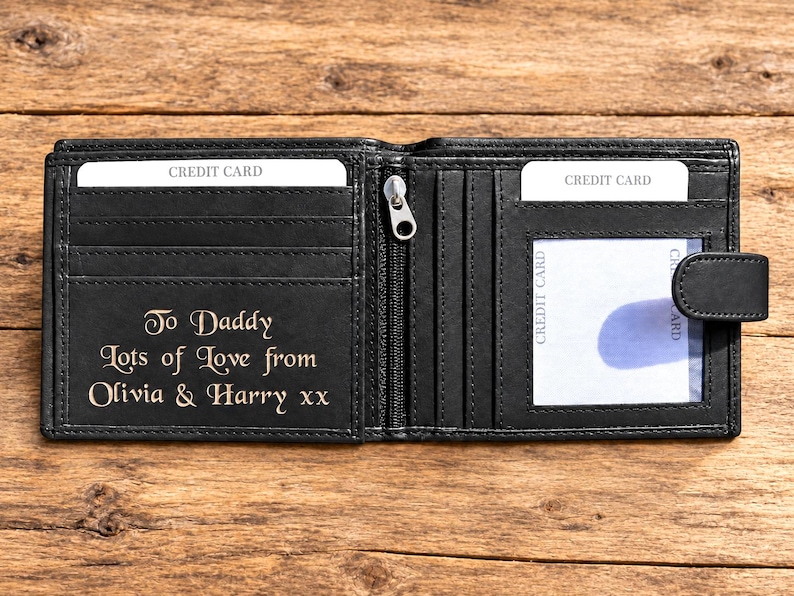 May include: A black bi-fold wallet featuring multiple card slots, a central zippered compartment, and a clear ID window. The interior is engraved with the text: To Daddy Lots of Love from Olivia & Harry xx. The wallet includes a snap button closure.
