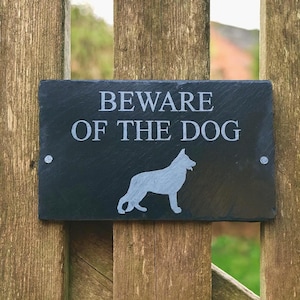 May include: A rectangular black slate sign with the words "BEWARE OF THE DOG" in white lettering. A white silhouette of a dog is below the text. The sign is mounted on a wooden fence.