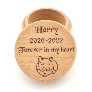 Może przedstawiać: Okrągłe pudełko z jasnego drewna ze zdejmowaną pokrywką. Na pokrywce wygrawerowano imię Harry, daty 2020-2022, napis Forever in my heart oraz rysunek liniowy małego chomika, wszystko wyśrodkowane na okrągłej powierzchni przedmiotu.
