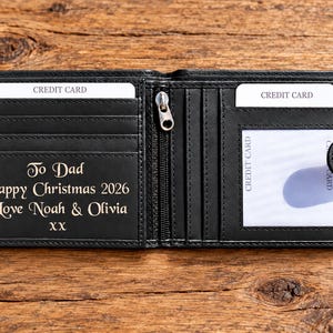 May include: A black bi-fold wallet featuring multiple credit card slots, a clear ID window, and a central zippered compartment. The interior is engraved with the text: To Dad, Happy Christmas 2026, Love Noah & Olivia, XX. The wallet includes a snap closure.