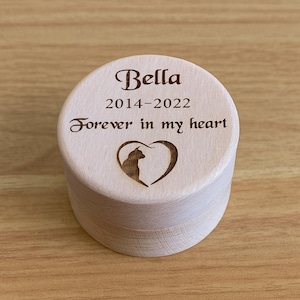 May include: A light-coloured wooden pet urn with the name "Bella", dates 2014-2022, and the phrase "Forever in my heart" engraved on the lid. A heart-shaped design with a cat silhouette is also featured.