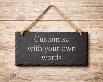 May include: A rectangular black slate sign with rough, uneven edges, suspended by a tan jute rope. The center of the slate features the engraved white text: Customise with your own words. The sign has a dark, textured, stone-like surface.