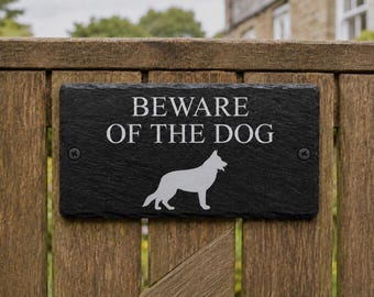 May include: A rectangular black slate sign featuring the engraved white text BEWARE OF THE DOG and a silhouette of a German Shepherd dog. The sign has two black screws on the left and right sides for mounting purposes.