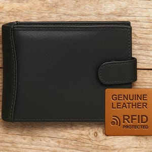 May include: Black leather wallet with a snap closure. The wallet has a stitched border and a brown leather tag that reads "GENUINE LEATHER" and "RFID PROTECTED". The wallet is on a wooden surface.