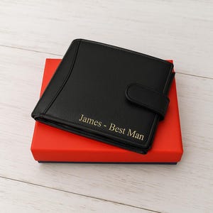 May include: A black leather wallet with a snap closure, resting on a red box. The wallet has the inscription "James - Best Man" in gold lettering. The box is rectangular and has a blue interior.