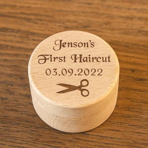 May include: A circular wooden box with a lid, featuring the engraved text "Jenson's First Haircut" and the date "03.09.2022", along with a scissors graphic. The box is made of light-colored wood and is designed to hold a keepsake.