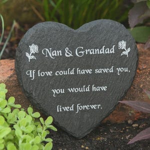 Op de afbeelding: Een hartvormige, donkergrijze leisteen gedenksteen met de tekst "Nan & Grandad" en een ontroerende boodschap in het wit. Bloemenaccenten flankeren de tekst, geschikt voor tuin of gedenkplaats.