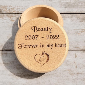 Op de afbeelding: Een ronde houten herinneringsdoos met het deksel iets open. Op het deksel staan de woorden "Beauty 2007 - 2022 Forever in my heart" en een paardenkop in een hart gegraveerd.