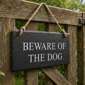 A dark grey rectangular slate sign hangs by a rustic twine rope from a weathered wooden garden gate, engraved with the words BEWARE OF THE DOG.