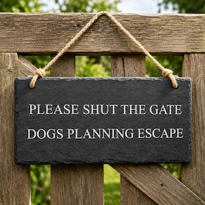 A dark grey rectangular slate sign hangs by a rope from a wooden gate, displaying the engraved text PLEASE SHUT THE GATE DOGS PLANNING ESCAPE.