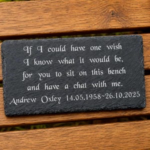 Può includere: Una targa commemorativa rettangolare in ardesia nera con bordi ruvidi e strutturati. La superficie è incisa con testo bianco che recita: If I could have one wish I know what it would be, for you to sit on this bench and have a chat with me. Andrew Oxley 14.05.1958~26.10.2025.