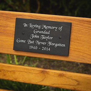 May include: A rectangular black slate memorial plaque with the inscription "In Loving Memory of Grandad John Taylor Gone But Never Forgotten 1940 - 2014." The plaque is mounted on a wooden bench.