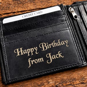 Open black leather wallet resting on a wooden surface with the engraved text "Happy Birthday from Jack" visible on the inside flap.