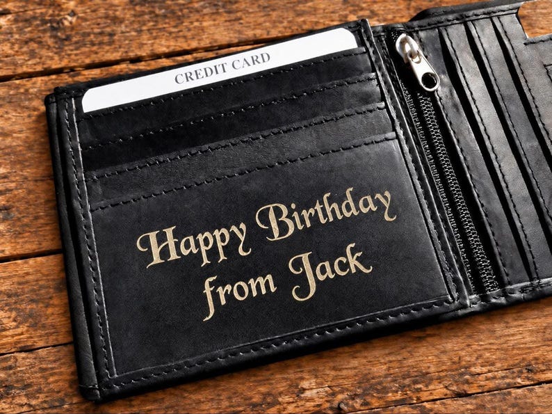 Open black leather wallet resting on a wooden surface with the engraved text "Happy Birthday from Jack" visible on the inside flap.
