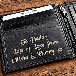 Open black leather wallet on a wooden surface, showing card slots and gold engraved text that reads To Daddy lots of love from Olivia & Harry xx.