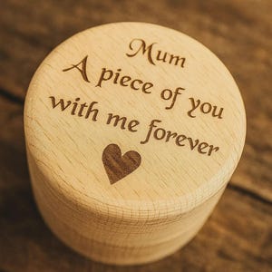 Puede incluir: Una caja de madera de color claro con tapa. La tapa está grabada con la palabra "Mum", la frase "A piece of you with me forever" y un símbolo de corazón. La caja es cilíndrica.