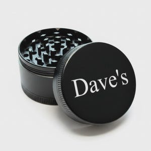 May include: A black, cylindrical herb grinder with the lid slightly ajar. The lid features the white text "Dave's". The grinder's interior has a silver, multi-pronged design. The exterior has a textured, ridged edge.