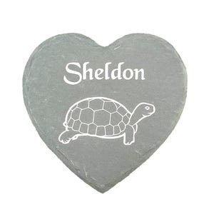 Pode incluir: Pedra de ardósia cinza em forma de coração com o nome "Sheldon" em escrita branca sobre um desenho de contorno branco de uma tartaruga. Um item decorativo.