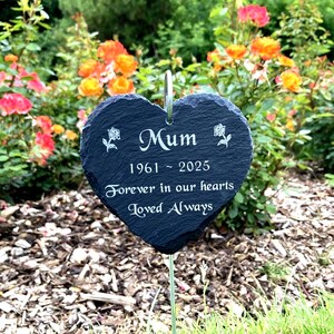 May include: A heart-shaped memorial garden stake made of dark gray slate. The stake reads "Mum 1961-2025 Forever in our hearts Loved Always" in white lettering. Two small rose illustrations flank the word "Mum". The stake is in a garden.