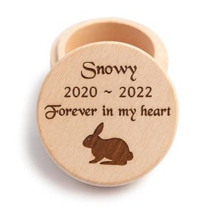 Könnte beinhalten: Kleine, offene Holzkiste mit rundem Deckel. Der Deckel ist mit dem Namen "Snowy", den Daten "2020 ~ 2022", dem Satz "Forever in my heart" und einer Kaninchensilhouette graviert. Die Kiste ist aus hellem Holz.
