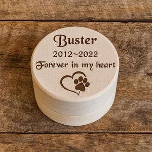 Könnte beinhalten: Eine runde, hellfarbene Holzkiste mit Deckel. Der Deckel ist mit dem Namen "Buster", den Daten "2012-2022" und dem Satz "Forever in my heart" sowie einem Herz- und Pfotenabdruck-Design graviert. Die Kiste steht auf einer Holzoberfläche.