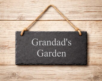 May include: A rectangular black slate sign with rough, uneven edges, featuring the engraved white text "Grandad's Garden" in a serif font. The sign is suspended by a thick, natural-colored jute rope attached to two metal eyelets.