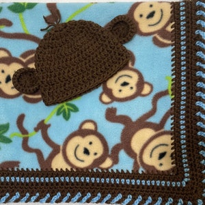 May include: A brown crocheted monkey hat with brown ears and a blue fleece blanket with a monkey print and a brown and blue crocheted border.