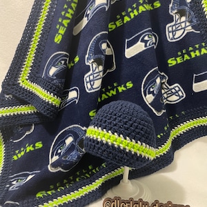 May include: A blue fleece baby blanket with a white and green crocheted border. The blanket features a repeated pattern of the Seattle Seahawks logo. A matching crocheted hat is displayed on top of the blanket.