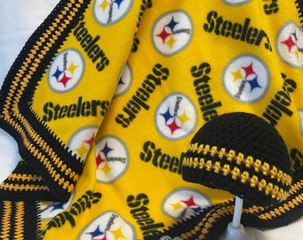 May include: A yellow and black Steelers-themed baby blanket and matching crochet hat. The blanket features repeating Steelers logos and text. The hat is black with yellow stripes. The blanket has a black and yellow crochet border.