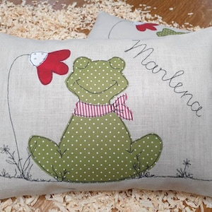 Op de afbeelding: Een lichtgrijs kussen met een groene kikker met rode stippen en een rood-wit gestreepte strik. De kikker zit op een groene bloem met een rode kern. Het kussen heeft de naam "Marlena" geborduurd.