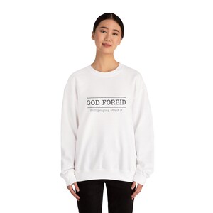 May include: White sweatshirt with the text "GOD FORBID" above the text "Still praying about it." in a dark blue font. The sweatshirt is a solid white color and has a crew neck.