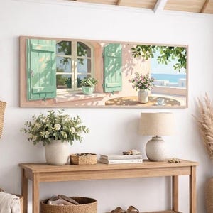 Può includere: Una stampa artistica rettangolare che raffigura una finestra con persiane verde chiaro affacciata su una scena costiera, esposta sopra una console in legno. Il tavolo ospita una lampada, un vaso di fiori e un cesto, con un cesto intrecciato e un cappello nelle vicinanze.