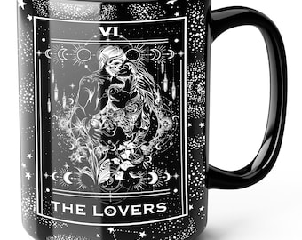 Puede incluir: Una taza de cerámica negra con un estampado gráfico blanco de un diseño de carta del tarot. La ilustración muestra dos figuras esqueléticas abrazadas, rodeadas de motivos celestiales, estrellas y el número romano VI. El texto THE LOVERS aparece.