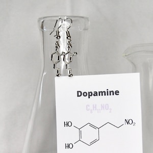 Op de afbeelding: Zilveren dopamine-molecuul oorbellen die hangen aan een glazen beker. De beker heeft een label met de tekst "Dopamine" en de chemische formule voor dopamine. De tekst onder de formule legt uit dat dopamine een neurotransmitter is die essentieel is voor motorische controle en de regulering van het beloningssysteem van de hersenen.