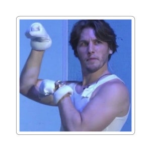 May include: A man wearing a white tank top flexes his bicep while wearing white gloves. The background is a light blue wall.