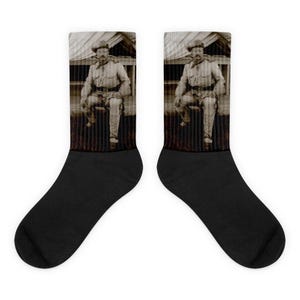 May include: Pair of black socks featuring a sepia-toned image of a man in a hat and uniform, seated in front of a wooden structure. The socks have a black foot and a sepia-toned upper.