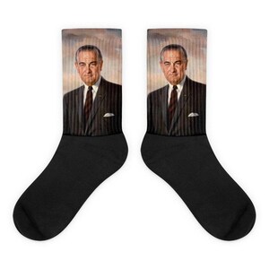 May include: Pair of black socks featuring a portrait of a man in a suit. The portrait is in color and shows the man from the chest up, with a neutral background. The socks are made of a soft material and are designed to fit most adult sizes.