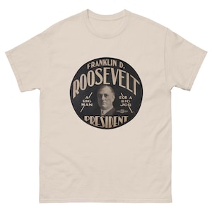 May include: A light beige t-shirt featuring a black and white graphic of Franklin D. Roosevelt. The graphic is a circular design with the name "ROOSEVELT" and the word "PRESIDENT" prominently displayed. The shirt has short sleeves and a crew neck.