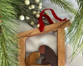 May include: A wooden nativity ornament featuring a light-toned frame and three stylized figures in varying shades of brown wood. The figures are arranged within the frame, suspended by a bright red velvet ribbon for hanging purposes.