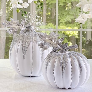 May include: Two white pumpkins with silver glitter and beaded accents. The pumpkins are sitting on a white surface.